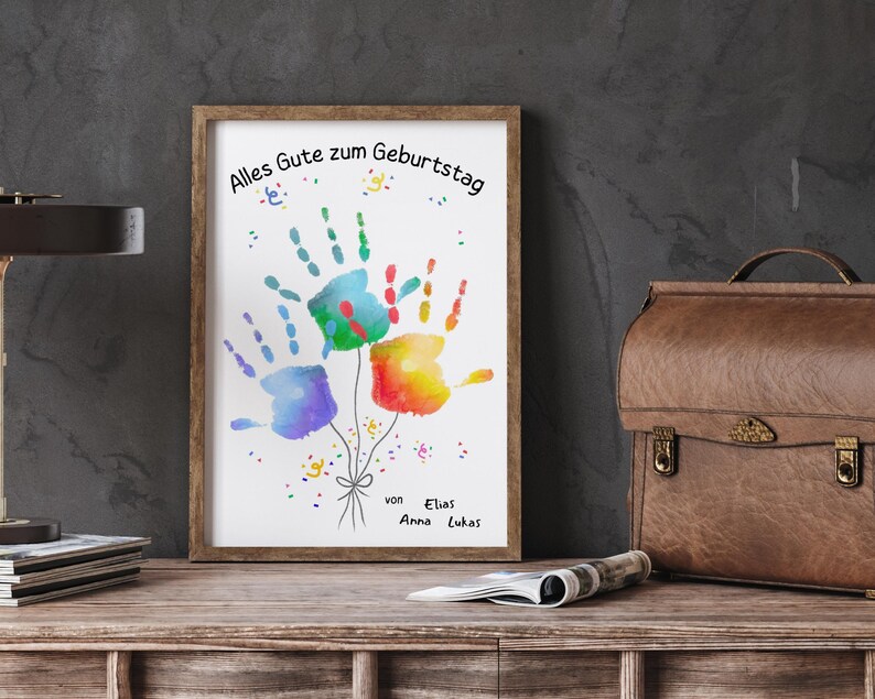 Peut inclure: Une impression encadr&eacute;e avec des empreintes de mains color&eacute;es en forme de ballons. L'&oelig;uvre comprend le texte allemand "Alles Gute zum Geburtstag" et les noms "Elias, Anna, Lukas". Les empreintes sont bleues, vertes, rouges et jaunes.