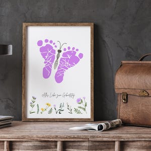 K&ouml;nnte beinhalten: Ein gerahmter Druck mit einem Schmetterlingsdesign aus lilafarbenen Fu&szlig;abdr&uuml;cken. Das Kunstwerk enth&auml;lt den Text "Alles Liebe zum Geburtstag" und florale Illustrationen unten. Der Rahmen ist braun.