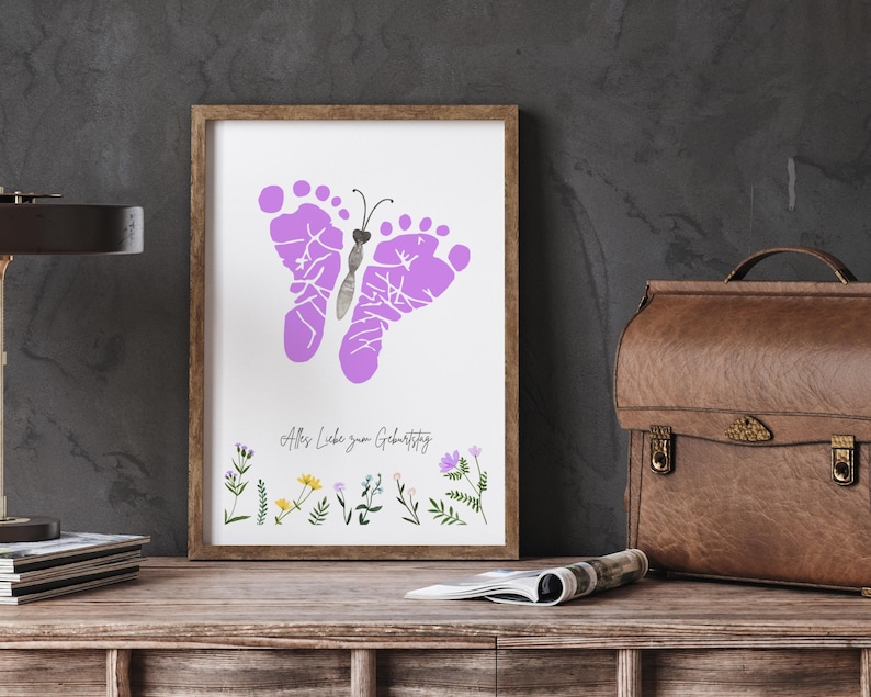 K&ouml;nnte beinhalten: Ein gerahmter Druck mit einem Schmetterlingsdesign aus lilafarbenen Fu&szlig;abdr&uuml;cken. Das Kunstwerk enth&auml;lt den Text "Alles Liebe zum Geburtstag" und florale Illustrationen unten. Der Rahmen ist braun.