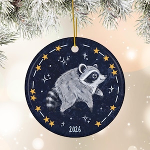 May include: A round ceramic ornament featuring a gray and white raccoon illustration centered on a dark blue background with yellow stars and white sparkles. The year 2026 is printed in white at the bottom, and a gold string is attached for hanging.