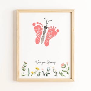 Peut inclure: Une œuvre d'art encadrée avec un motif de papillon fait d'empreintes de pieds de bébé roses. Le papillon a un corps gris et des antennes noires. Les mots "I love you, Grammy" sont écrits en dessous, avec des accents floraux.
