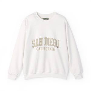 Może przedstawiać: Biała bluza z długim rękawem i okrągłym dekoltem, z napisem SAN DIEGO CALIFORNIA wykonanym wypukłą, beżową czcionką na klatce piersiowej. Odzież posiada prążkowany kołnierzyk, mankiety i dół, a wewnątrz znajduje się mała metka z napisem ENOXWEAR.