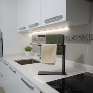 Pode incluir: Uma cozinha moderna com um porta-toalhas preto que segura duas toalhas, uma bege e outra verde. O porta-toalhas está sobre uma bancada branca ao lado de uma placa de fogão preta. Armários brancos e uma pia são visíveis no fundo.