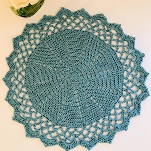 May include: A teal blue crocheted doily with a delicate lace border. The doily is round and has a center that is slightly raised.