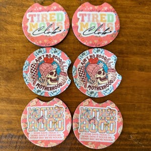 May include: Set of six car coasters with floral patterns and various text designs. Two coasters read "TIRED MOMS Club" in colorful letters. Two feature a skull with a beanie and the text "AIN'T NO HOOD Motherhood". The last two say "MOTHERHOOD" with text below.