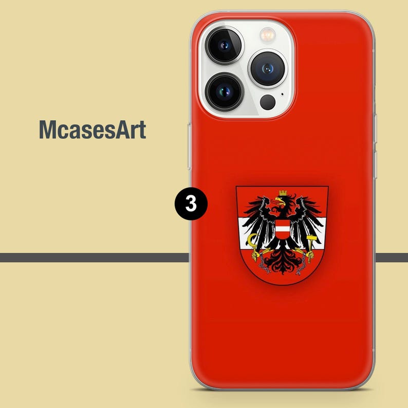 Peut inclure: Une coque de t&eacute;l&eacute;phone rouge pour iPhone avec un blason blanc et rouge repr&eacute;sentant un aigle noir avec une couronne dor&eacute;e. La coque a un prot&egrave;ge-objectif d'appareil photo transparent.