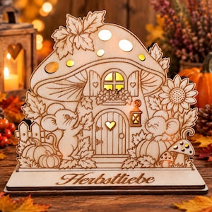 Könnte beinhalten: Eine dekorative Lampe aus hellem Holz in Form eines Pilzhauses mit Ausschnitten von zwei Mäusen, Kürbissen, Blättern und einer Sonnenblume. Der Sockel trägt den eingravierten Text "Herbstliebe". Die Struktur ist von innen beleuchtet.