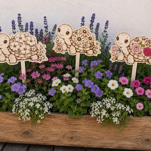 Puede incluir: Tres estacas de jardín de madera con ilustraciones de tortugas grabadas con láser y caparazones florales. Las tortugas están colocadas en una jardinera rectangular de madera llena de lavanda, flores rosas y pequeñas flores blancas.