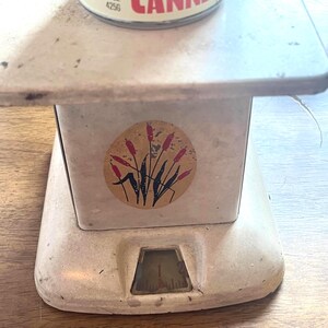 May include: An antique cream-colored kitchen scale with a round sticker featuring cattails. A can of food is on the scale. The can reads "Canning" and "Net Wt 15 oz". The scale has a dial at the bottom.