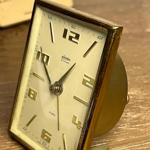 May include: A vintage gold-toned alarm clock with a rectangular face. The clock face is cream-colored with black numerals and hands. The brand name "Linden Blackforest" is printed on the face. The clock has a small alarm setting dial on the back.