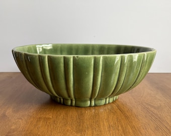 May include: A green ceramic bowl with a ribbed exterior. The bowl has a wide, oval shape and sits on a small, rounded base. The bowl is a solid, muted green color and appears to be vintage.