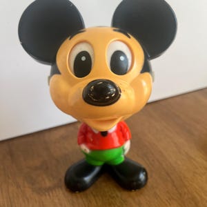 May include: A vintage plastic Mickey Mouse figurine with black ears, yellow face, and red and green clothing.