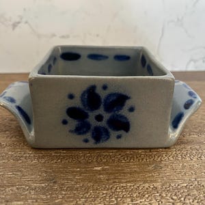 May include: A small, rectangular, stoneware dish with a light gray base and blue floral design. The dish has two small handles and is decorated with blue dots and a large flower. The dish is sitting on a wooden surface.