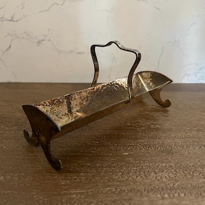 May include: A vintage silver-toned metal serving dish with a handle and curved legs. The dish has a rectangular shape with a hammered texture and a decorative handle. The piece is likely used for serving small items.