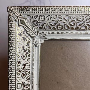 May include: A rectangular picture frame featuring an ornate metal border with a distressed white finish over a gold-toned base. The frame displays intricate cut-out floral and leaf patterns along the edges with a decorative corner accent piece.