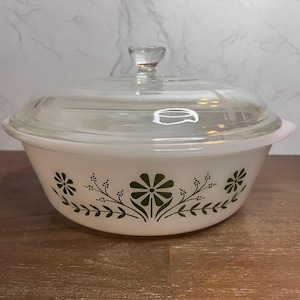 May include: A round white glass casserole dish with a clear glass lid and a knob handle. The exterior features a repeating green floral pattern consisting of stylized daisies and leafy sprigs encircling the base of the bowl.