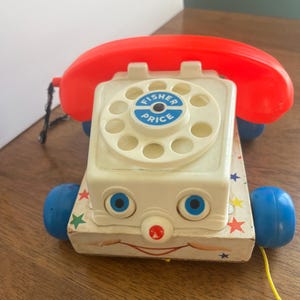 May include: Vintage Fisher-Price toy phone with a red receiver and a cream-colored body. The toy has blue eyes, a red nose, and a smiling mouth. The phone sits on a wooden base with blue wheels and a pull string. The dial has the Fisher-Price logo.