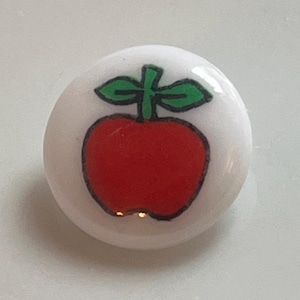 May include: A round, white button with a glossy finish. The button features a red apple design with a black outline and green leaves. The apple is centered on the button.