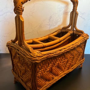 May include: A woven rattan wine bottle carrier with a handle. The basket is rectangular with a woven pattern and has a handle that arches over the top. The interior has dividers to hold bottles. The basket is a warm brown color.