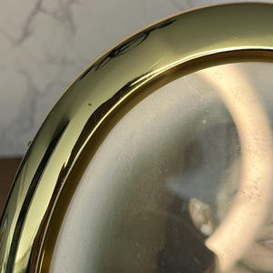 May include: Close-up of a gold-colored oval picture frame. The frame has a shiny, reflective surface and appears to be made of metal. The photo inside is slightly visible, showing a person in a dark outfit.
