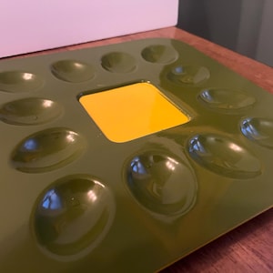 May include: Olive green paint palette with a glossy finish. The palette features 12 oval wells and a central square well in bright yellow. The palette is designed for mixing paints and is made of a durable material.