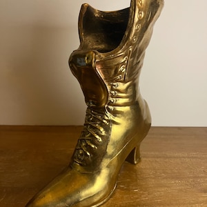 May include: A gold-colored decorative boot with a pointed toe and a small heel. The boot features a lace-up design and button details along the side. The boot is made of a shiny, metallic material and is likely a decorative item.
