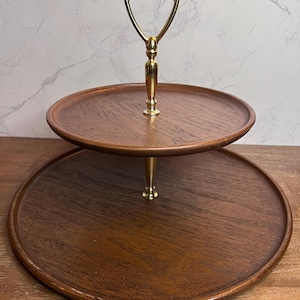 May include: A two-tiered serving stand featuring two circular wooden trays with a visible grain pattern. The trays are connected by a central gold-toned metal rod that includes a decorative handle at the top for carrying and support.