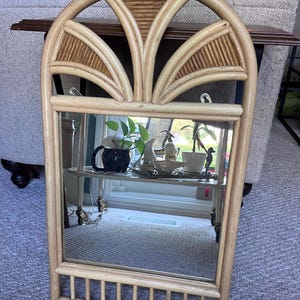 May include: A rectangular wall mirror featuring a light brown natural rattan frame. The top section has an arched design with decorative fan-shaped rattan slats, while the bottom section includes a series of vertical rattan bars.