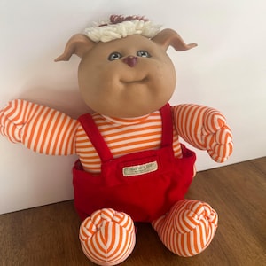 May include: A brown plush toy with a white and red striped shirt and red overalls. The toy has a white and red hat with a brown pom-pom on top. The toy has a smiling face with brown eyes and a pink nose. The toy is sitting on a wooden surface. The toy has a tag that says "Cabbage Patch Kids" on it.
