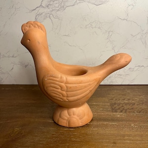 May include: A terracotta chicken-shaped candle holder. The bird has a detailed comb and tail feathers. The candle holder is a light orange-brown color and sits on a round base with a floral design. The candle holder is made of ceramic.