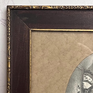 May include: An antique framed print with a dark brown wooden frame and gold trim. The print is set against a tan background and features a black and white image of a building.