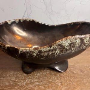 May include: An oval-shaped ceramic bowl featuring a dark brown interior and exterior glaze. The rim is accented with a mottled, light cream-colored drip pattern that extends down the sides. The bowl is supported by a solid, dark brown pedestal base.