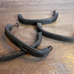 May include: A set of three dark, weathered metal drawer pulls with a curved, arch-shaped design. Each handle features a smooth, matte finish with visible signs of age and wear, and includes two small, threaded metal screws attached at the ends.