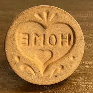 May include: A round, light brown ceramic knob with the word "HOME" embossed inside a heart shape. The knob has a rustic, handcrafted appearance, suitable for furniture or decorative projects.