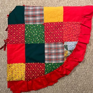 May include: A patchwork quilt with a variety of fabrics in red, green, yellow, and plaid patterns. The quilt has a red ruffled border and red ties on one corner. The quilt is likely handmade and has a vintage aesthetic.