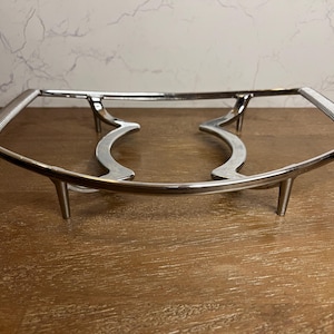 May include: A silver-colored metal serving stand with a rectangular frame and curved supports. The stand has a ribbed design on the handles and is elevated by four short legs. The stand is designed to hold a dish.