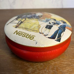 May include: A vintage red and white tin with a lid featuring a colorful illustration of children playing with hoops and an older woman. The word "Nestle" is printed in gold on the lid.