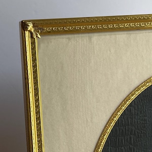 May include: Close-up of a framed artwork with a gold-colored ornate frame. The artwork features a black, textured oval shape against a cream-colored background. The frame has decorative detailing.