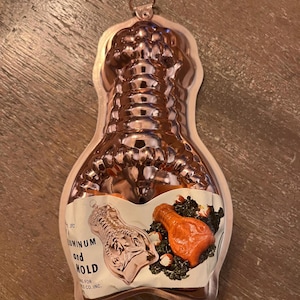 May include: A copper-colored metal lobster-shaped mold with a hanging loop at the top. A paper label is attached to the front, featuring an illustration of the mold and a cooked lobster, with text that reads Aluminum and Mold.