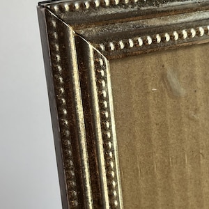 May include: Close-up of a rectangular picture frame with a gold-colored, ornate border. The frame has a textured surface with a series of small, raised dots along the edges. The frame's corner is visible, showing the detailed craftsmanship.