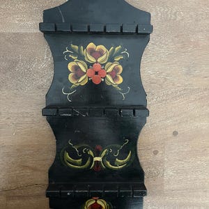 May include: Black wooden wall shelf with three tiers, adorned with hand-painted floral designs in yellow, red, and green. The shelf has a decorative top with a hole for hanging. The floral patterns are symmetrical and stylized.
