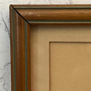 May include: A corner view of a framed artwork. The frame is made of wood with a brown finish and a thin green accent. The artwork is behind a cream-colored mat.