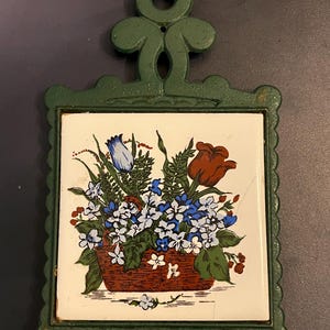 May include: Green cast iron trivet with a floral tile insert. The tile features a basket of flowers in red, blue, and white. The trivet has a decorative handle and scalloped edges.