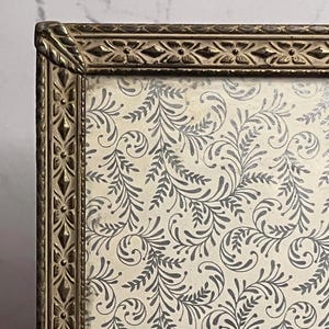 May include: An antique gold-toned picture frame with ornate detailing. The frame surrounds a cream-colored background with a repeating pattern of gray, stylized floral and leaf designs. The frame's edges have a textured, decorative pattern.