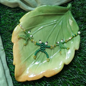 May include: A beaded necklace with three green frog charms on a green leaf-shaped plate. The frogs are made of green and white beads.