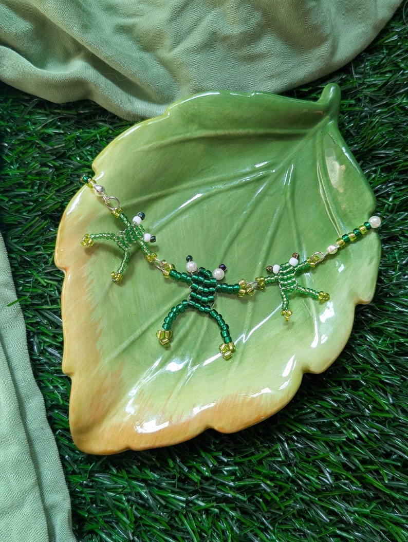 May include: A beaded necklace with three green frog charms on a green leaf-shaped plate. The frogs are made of green and white beads.
