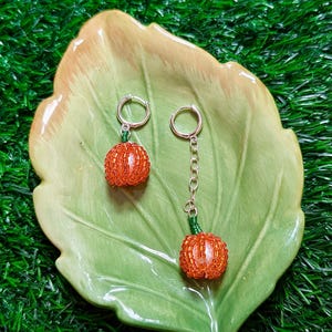Op de afbeelding: Een paar oranje kralen pompoen oorbellen met zilveren hoepels en groene stelen. Eén oorbel is een simpele hoepel, terwijl de andere een ketting heeft. De oorbellen worden gepresenteerd op een groen blad-vormig schaaltje.