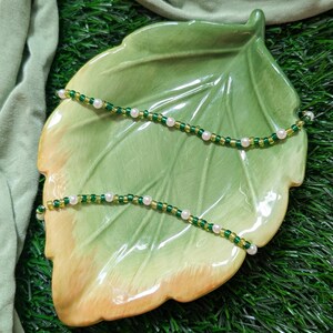 May include: Two beaded necklaces with green and white beads on a green leaf-shaped plate. The plate is on a green surface.