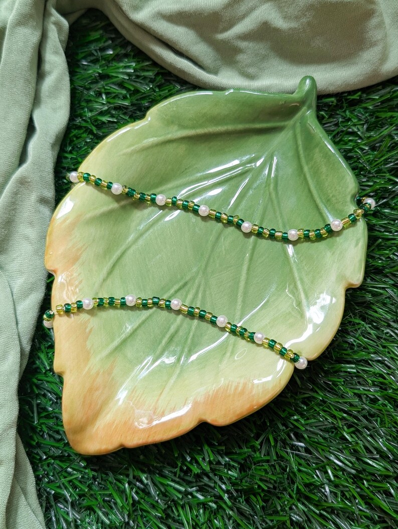 May include: Two beaded necklaces with green and white beads on a green leaf-shaped plate. The plate is on a green surface.