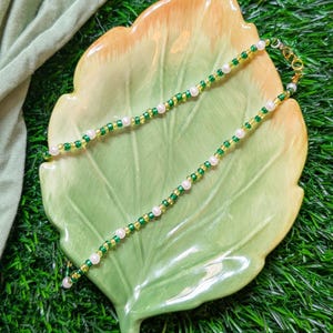 May include: A delicate gold chain necklace with alternating green and white beads. The necklace is displayed on a green leaf-shaped plate.